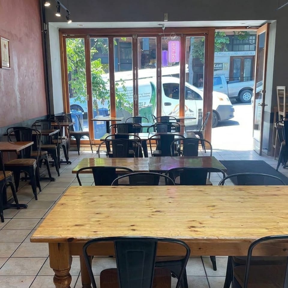 Empty restaurant dining room with wooden tables, black chairs, artwork, and sunlight streaming through front windows.